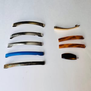 May include: A selection of hair barrettes in different colours and designs. The barrettes include gold, silver, blue, and tortoiseshell patterns. The hair accessories are made of metal and plastic. The barrettes are arranged on a white surface.
