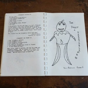 May include: Open cookbook pages with handwritten recipes for Strawberry Chiffon Pie and Strawberry Ice Cream Pie. A whimsical drawing of a fruit and vegetable man is on the right page. The text includes ingredients and instructions.