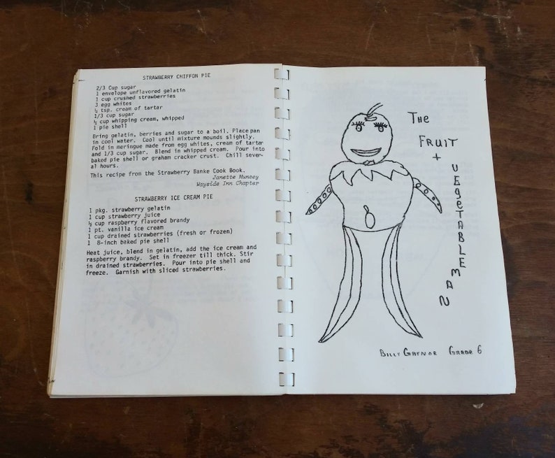 May include: Open cookbook pages with handwritten recipes for Strawberry Chiffon Pie and Strawberry Ice Cream Pie. A whimsical drawing of a fruit and vegetable man is on the right page. The text includes ingredients and instructions.