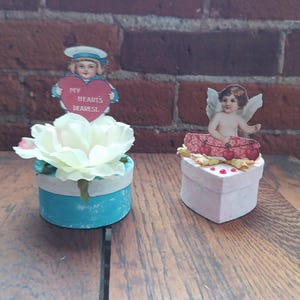 May include: Two decorative Valentine's Day themed items. One is a blue and white painted pot with a white flower and a vintage image of a child holding a heart that says "My Heart's Dearest." The other is a pink heart-shaped box with a cherub image.