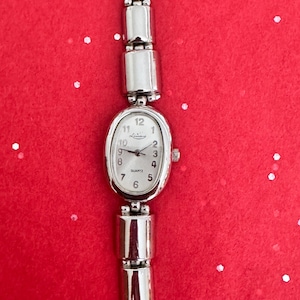 May include: Silver-toned quartz watch with an oval face and a silver band. The watch face has a white background with black numerals and hands. The watch is on a red background.
