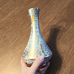 Puede incluir: Un jarrón de cerámica azul claro con detalles dorados. El jarrón tiene una superficie texturizada y un cuello estrecho.