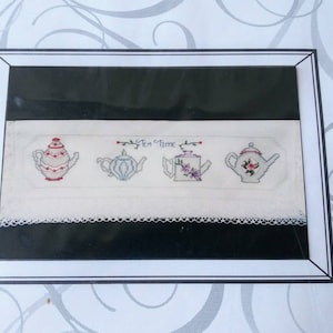 May include: A white tea towel with a lace trim and four embroidered teapots. The teapots are decorated with flowers and the text "Tea Time" is embroidered in the center.