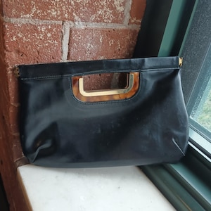 May include: A black clutch bag featuring a smooth, solid-colored exterior and a rectangular cutout handle. The handle is accented with a brown and gold tortoiseshell-patterned trim, providing a distinct contrast to the plain black body of the bag.