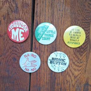 May include: Five round buttons with various phrases and graphics. One red button says "EXCUUUSSE ME" in white. Another green button says "USE YOUR HEAD LITTLE THINGS COUNT". A yellow button says "TO ERR IS HUMAN TO REALLY MESS UP TAKES A ABTU9MOJ". A white button says "NA-NO NA-NO" with a hand graphic. A white button says "BIONIC BUTTON".