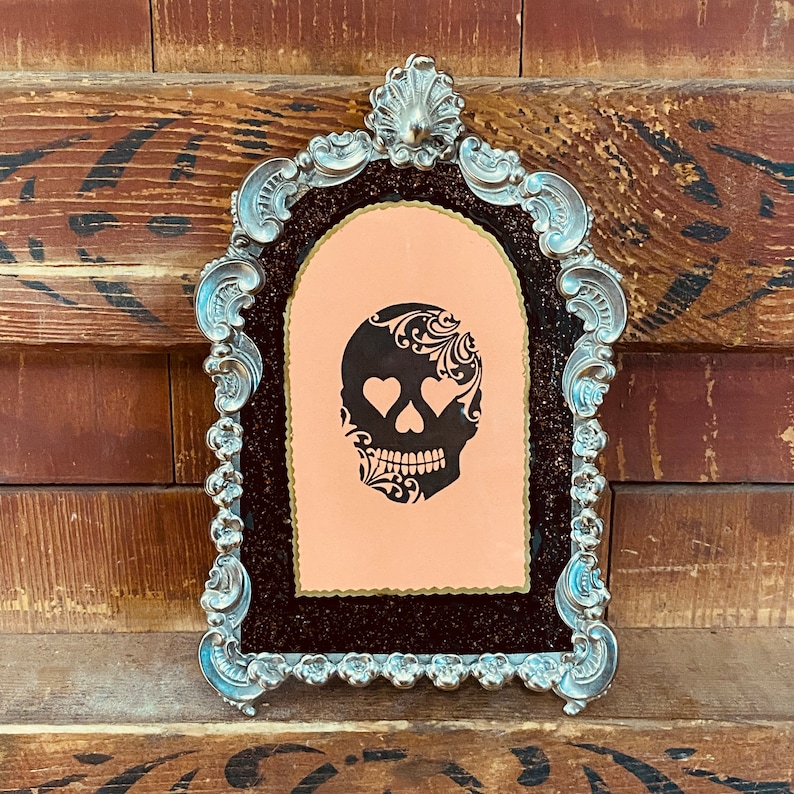 May include: A silver ornate frame with a black background and a pink mat. The frame features a black and white illustration of a skull with hearts for eyes.
