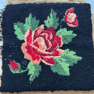May include: A square, handmade rug with a floral design. The rug features a large red and peach rose with green leaves against a black background. The edges are fringed with a tan fabric.