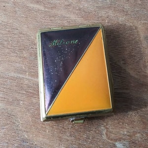 Peut inclure: Un compact vintage avec un design noir et orange. Le compact a une bordure dorée et le mot "Malrone" est écrit sur le devant.