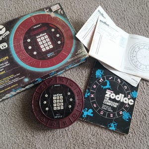 Könnte beinhalten: Ein Vintage Coleco Zodiac Astrology Computer Spielset bestehend aus einem runden kastanienbraunen elektronischen Gerät mit Ziffernblock, einem schwarzen Karton mit weißem Text, einer schwarzen Anleitung mit Tierkreiszeichen und einem weißen Papierdiagramm.