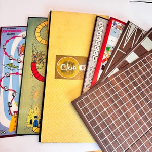 May include: A collection of vintage board games, including a yellow "Clue" game box with a gold emblem, and a "Scrabble" game. Other game boards feature colorful illustrations and grid patterns. The games are arranged in a fan shape.
