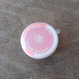 Könnte beinhalten: Ein runder, rosafarbener Rouge-Kompakt mit einem weißen Deckel. Der Deckel trägt den Text "CORAL BLUSH" und "VIVIANE WOODARD". Der Kompakt hat ein weißes "W"-Logo in der Mitte.