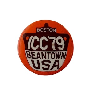 May include: Round orange button with the text "BOSTON" at the top. Below is a brown and white graphic with "(CC'79" and "BEANTOWN USA" in white text. The button has a small hole at the top.
