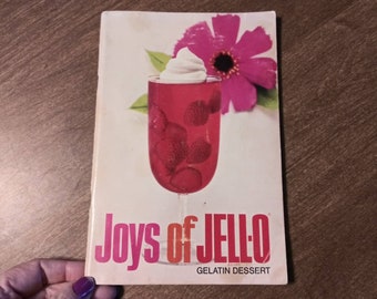 Op de afbeelding: Een vintage kookboek met de titel "Joys of JELL-O Gelatin Dessert" met een roze bloem en een glas rode gelatine-dessert met slagroom.