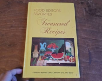 May include: A yellow cookbook with the title "Food Editors' Favorites Treasured Recipes" and a painting of fruit on the cover. The book is edited by Barbara Gibbs Ostmann and Jane Baker.