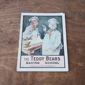 May include: Vintage cookbook cover featuring two teddy bears dressed as bakers. The cover is white with pink text that reads "The Teddy Bears Baking School. Compliments of The Fleischmann Co."