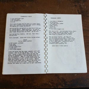 May include: Open cookbook pages with handwritten recipes for Strawberries Teresa and Thumbprint Cookies. The recipes are in black ink on aged, cream-colored paper. The book is open on a wooden surface.