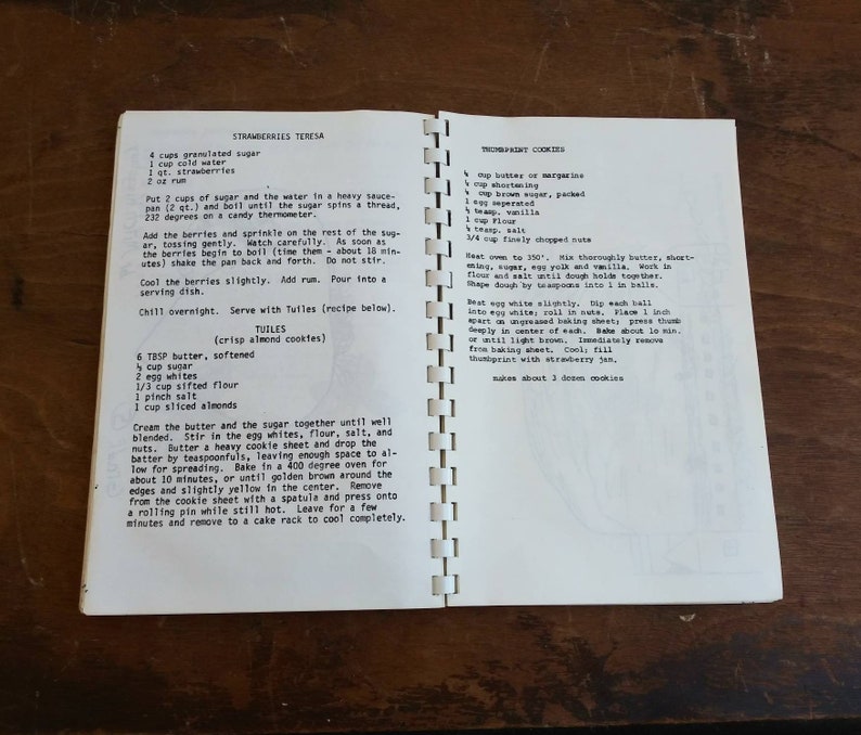May include: Open cookbook pages with handwritten recipes for Strawberries Teresa and Thumbprint Cookies. The recipes are in black ink on aged, cream-colored paper. The book is open on a wooden surface.