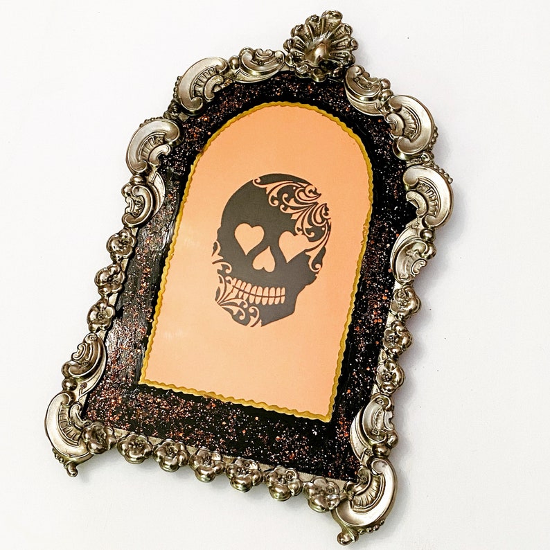 May include: A silver ornate frame with a black and orange glitter background. The frame has a cutout of a sugar skull with hearts for eyes.