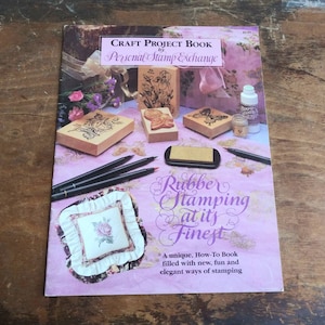 May include: A book titled "Rubber Stamping at its Finest" by Personal Stamp Exchange. The book cover features a variety of rubber stamps, ink pads, and craft supplies. The book is a how-to guide for rubber stamping.