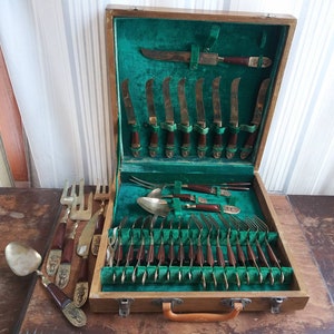 May include: A wooden box with a green velvet lining contains a set of brass silverware. The set includes knives, forks, and spoons. The box has a brass latch and a handle.
