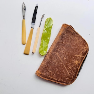 May include: An assortment of vintage manicure tools, including two knives, a cuticle pusher, and a green, leaf-shaped tool. A brown leather zippered pouch is also included. The tools have cream-colored handles and metal blades.