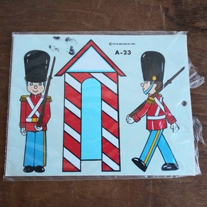 May include: A vintage sheet of paper decals featuring two toy soldiers in red and blue uniforms with black hats and a red and white striped candy cane house. The sheet is labeled "A-23" and "1978 DECOCAL INC."