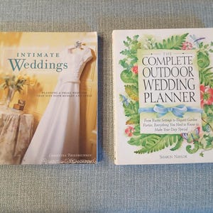 以下が含まれることがあります： 2冊のウェディングプランニング本が水色の表面に表示されています。左の本は「Intimate Weddings」というタイトルで、白いウェディングドレスが描かれています。右の本は、花のイラストが描かれた「The Complete Outdoor Wedding Planner」というタイトルです。