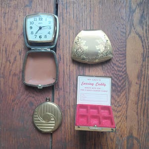 May include: An assortment of vintage accessories, including a travel clock, a gold-toned compact with floral detailing, a round compact, and an earring caddy. The earring caddy is open, revealing a red velvet interior. The clock shows the time.