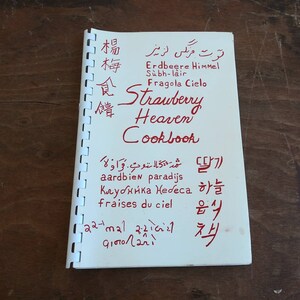 May include: A spiral-bound cookbook titled "Strawberry Heaven Cookbook" in red ink. The cover features text in multiple languages, including English, German, Italian, and Korean, with a wooden background.
