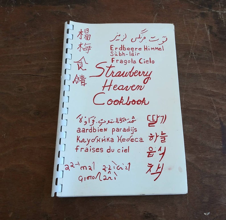 May include: A spiral-bound cookbook titled "Strawberry Heaven Cookbook" in red ink. The cover features text in multiple languages, including English, German, Italian, and Korean, with a wooden background.