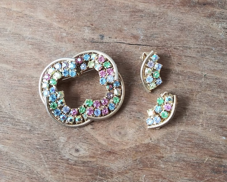 May include: A set of two gold-tone brooches with a heart-shaped design. The brooches are adorned with colorful rhinestones in shades of blue, green, pink, and yellow.