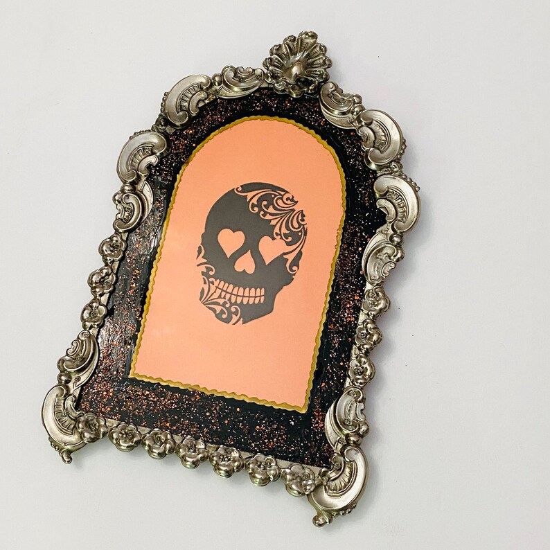 May include: A silver ornate frame with a black and gold glitter background. The frame has a peach-colored insert with a black and white skull design with hearts for eyes.