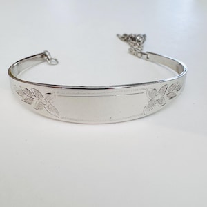 May include: A silver-toned bracelet with floral and leaf designs. The bracelet has a rectangular center section with engraved details and a chain extension. The bracelet is on a white surface.