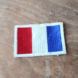 May include: A small, embroidered patch featuring the French flag. The patch is white with a red stripe on the left and a blue stripe on the right.