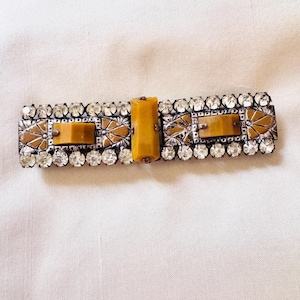 May include: A rectangular hair barrette featuring a central vertical amber-colored resin bar flanked by two horizontal amber-colored resin pieces. The piece is bordered by rows of clear round rhinestones set in a silver-toned metal frame.