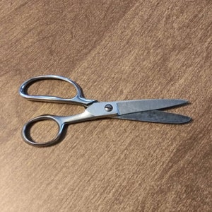 May include: A pair of silver scissors with a small, round handle and a larger, oval handle. The scissors are open and the blades are sharp.