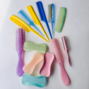 May include: A collection of colorful hairbrushes and combs in various shapes and sizes. The assortment includes yellow, blue, and green combs, along with pink and purple brushes. The items are arranged on a white surface.