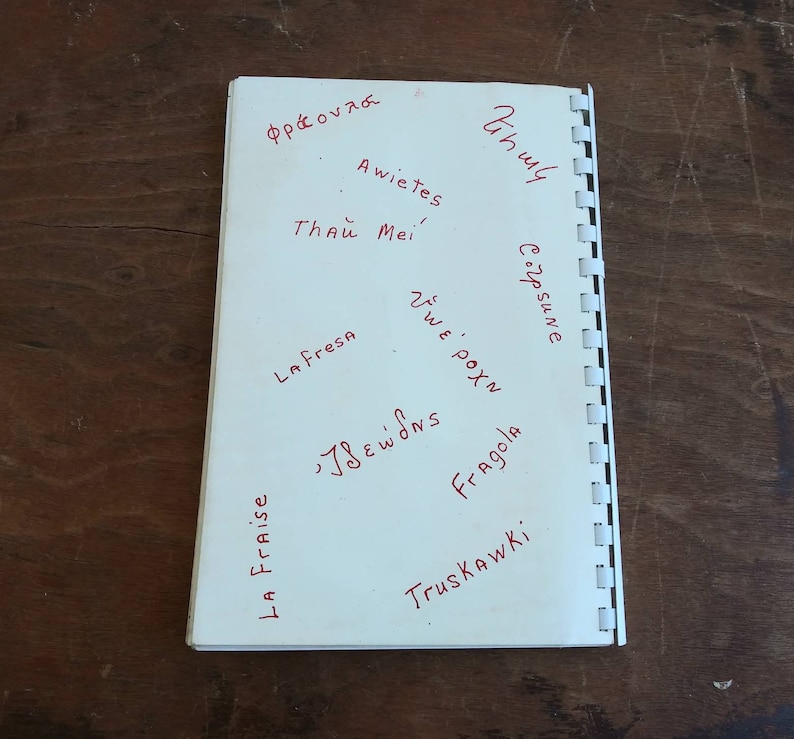May include: A white notebook with red handwritten words in various languages, including "La Fresa" and "Fragola". The notebook is open, revealing the pages and the spiral binding on the left side. The background is a dark wooden surface.