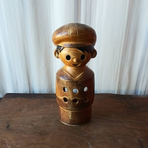 May include: A ceramic figurine of a person wearing a brown cap and a brown and tan patterned shirt. The figurine has a smiling face with black eyes and a black mouth.