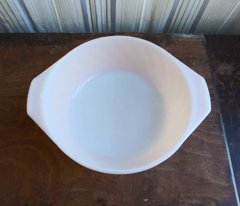 May include: A white, round baking dish with two handles. The dish is made of glass and has a slight pink tint.