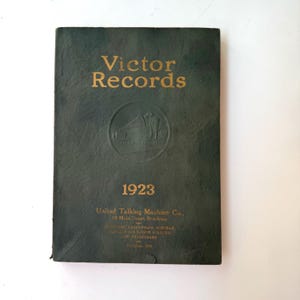 Puede incluir: Un libro verde con letras doradas que dice "Victor Records" y "1923". El libro es un catálogo de la United Talking Machine Co. ubicada en 19 Main Street, Braukion.