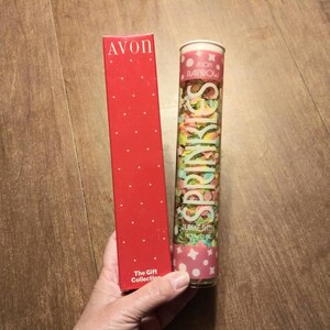 Puede incluir: Una caja roja con lunares blancos y la palabra "AVON" en letras blancas. La caja dice "The Gift Collection" en la parte inferior. Junto a la caja hay un tubo de plástico transparente lleno de confeti de colores. El tubo tiene una etiqueta rosa y blanca que dice "AVON RAINBOW" y "SPRINKLES" en una fuente blanca y cursiva. La etiqueta también dice "BUBBLE BATH" y "NET WT 1.5 OZ".