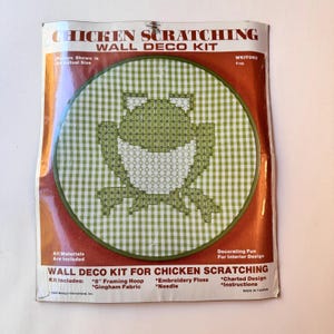 May include: A Chicken Scratching Wall Deco Kit with a frog design. The kit includes a 20 cm framing hoop, gingham fabric, embroidery floss, a needle, a charted design, and instructions. The frog is green and white.