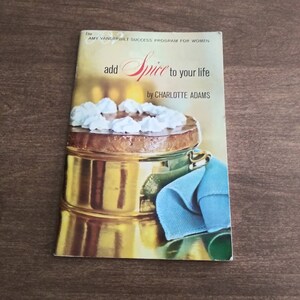 May include: A vintage cookbook titled "add Spice to your life" by Charlotte Adams. The cover features a golden cake dish with a cake topped with whipped cream and a blue napkin.