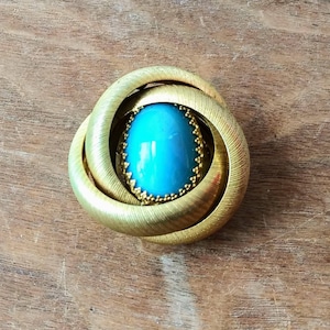 Puede incluir: Un anillo dorado con una piedra ovalada azul engastada en el centro. El anillo está diseñado con un patrón de nudo.