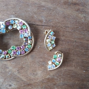 May include: A set of vintage jewelry featuring a gold-toned brooch and a pair of earrings. The brooch is shaped like a circle and is decorated with colorful rhinestones. The earrings are also decorated with colorful rhinestones.