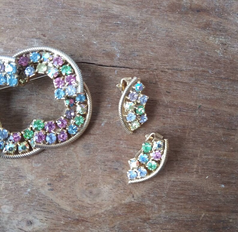 May include: A set of vintage jewelry featuring a gold-toned brooch and a pair of earrings. The brooch is shaped like a circle and is decorated with colorful rhinestones. The earrings are also decorated with colorful rhinestones.