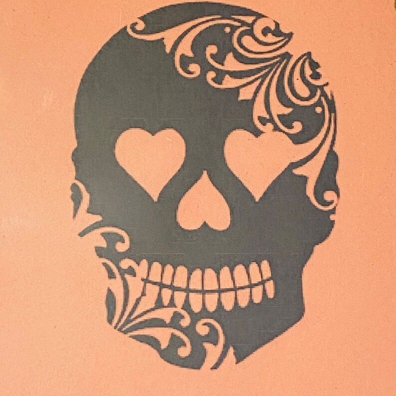 May include: Black and white silhouette of a skull with ornate floral details. The skull has two heart-shaped eyes and a toothy grin.