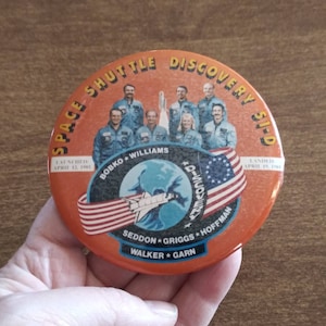 May include: A round orange button with a picture of the Space Shuttle Discovery 51-D. The button has the text "SPACE SHUTTLE DISCOVERY 51-D" around the edge. The button also has the text "LAUNCHED APRIL 12, 1985" and "LANDED APRIL 19, 1985".