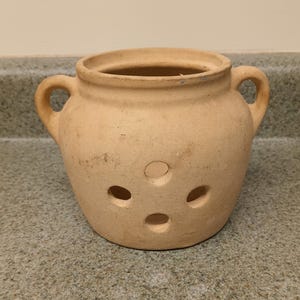 May include: A light tan, unglazed terracotta garlic keeper with a rounded body, two side handles, and four circular ventilation holes arranged in a diamond pattern on the front. The vessel has a wide, open top rim and a smooth, matte finish.